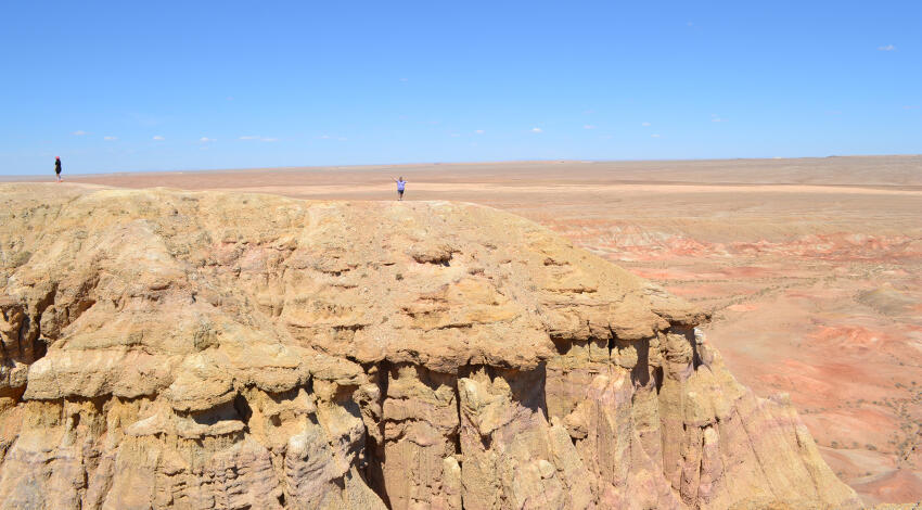 Two tourists standing on the edge of White Stupa, a dramatic sandstone cliff formation in the Gobi Desert under a blue sky.