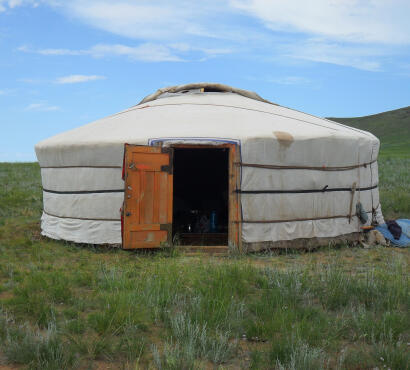 A
fully assembled traditional Mongolian ger covered in white canvas with an orange wooden door open, on a green grassy
steppe.