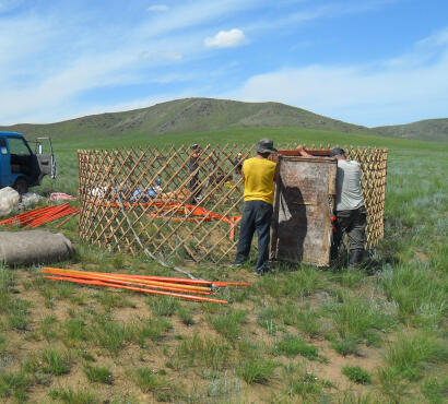 Three men arranging wooden lattice wall sections into a circle and fitting them to a door
                frame to assemble a Mongolian ger.