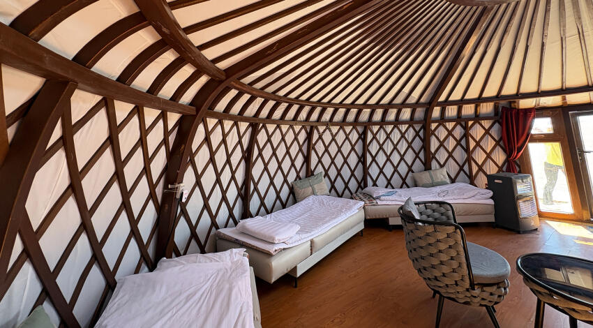 Three single beds with white bedding along the lattice wall of a ger, with a woven chair, table, and heater near the entrance.