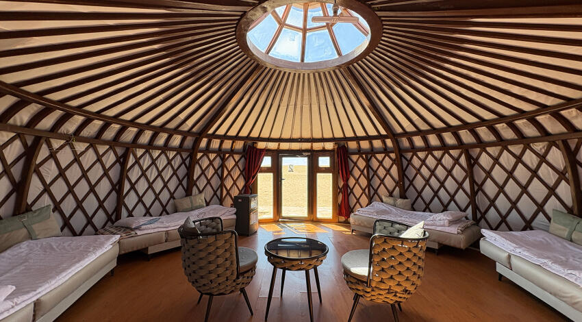 Single beds along the lattice walls of a Mongolian ger, two woven chairs, a small side table, and a glazed entrance door.