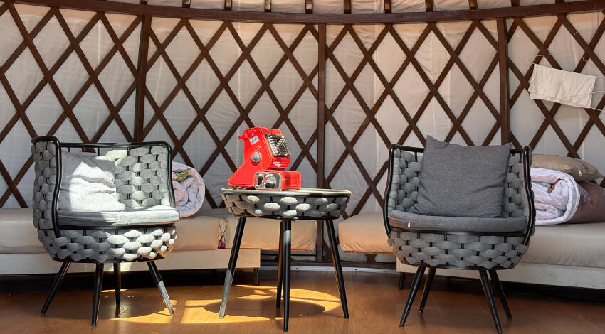 Two woven armchairs flanking a small round table inside a Mongolian yurt, with a red portable gas heater placed on the table.