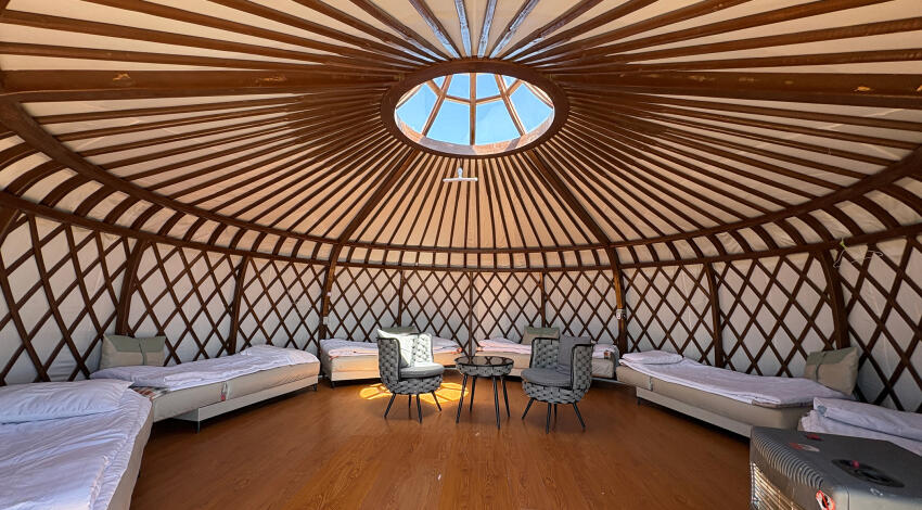 Yurt interior with a circular skylight, beds along the curved lattice walls, two woven chairs and a table in the center.